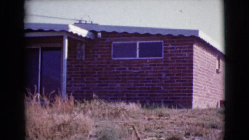1964 Cottonwood, Arizona 8mm view of the brick house, showing a dark opening at left, high windows, corrugated roof, and scrubby grass.
