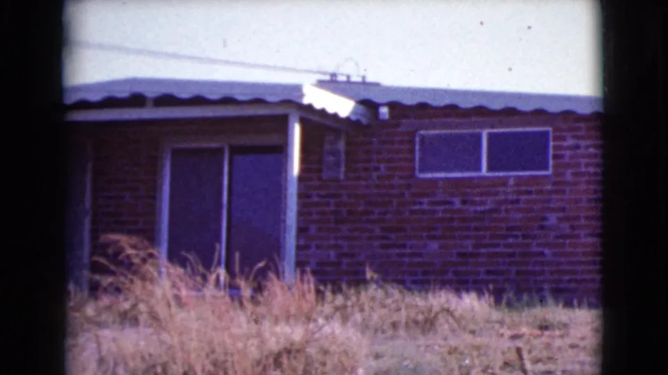 1964 Cottonwood, Arizona frame showing the house front more fully, with a doorway or patio opening, two windows, brick wall, and dry grass.
