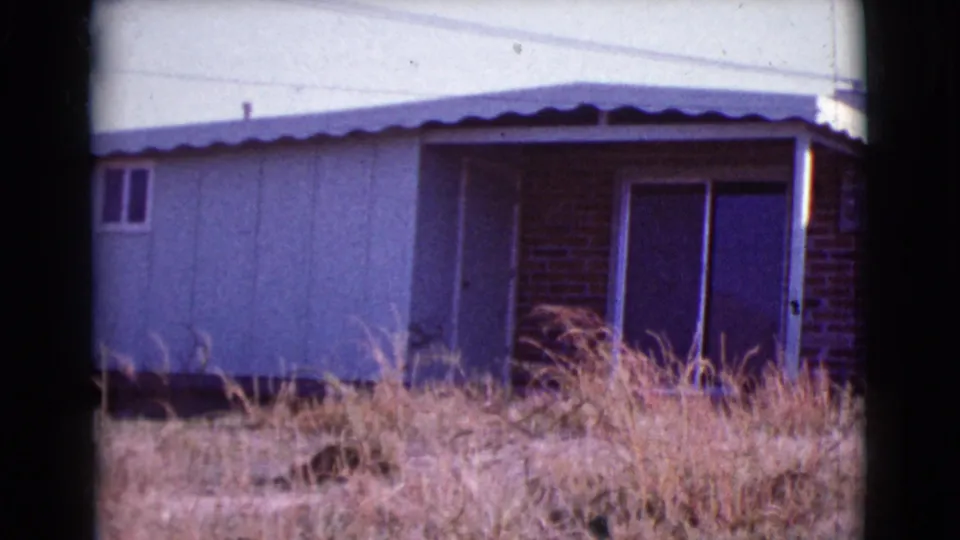 The frame shifts to a side angle of the Cottonwood house, showing pale siding, a brick section, sliding doors, small windows, and dry grass.