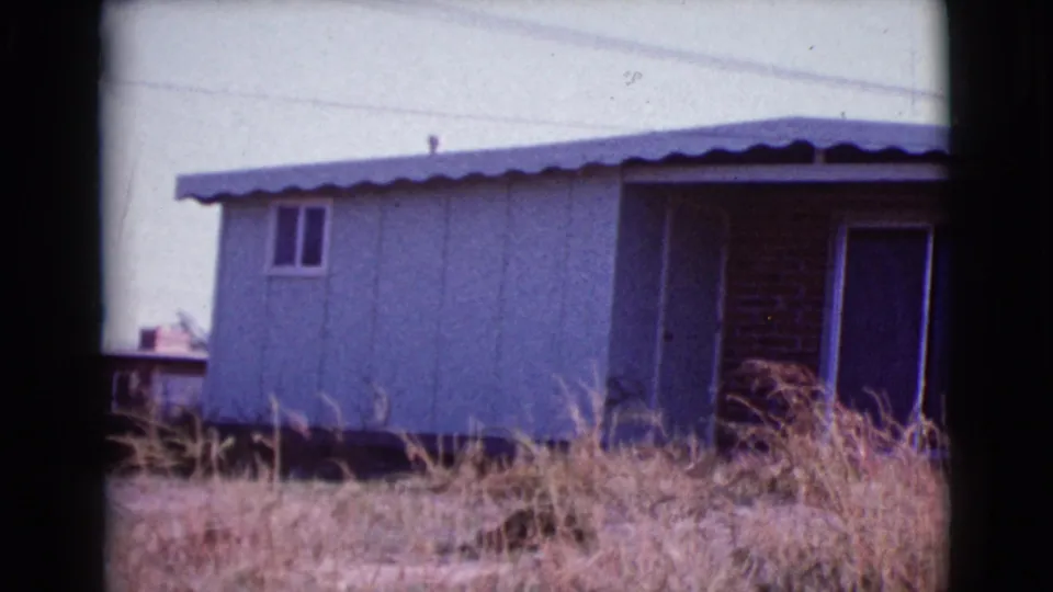 Final 1964 Cottonwood frame of the house corner, with pale siding, porch opening, sliding door, dry grass, and a glimpse of nearby structures.