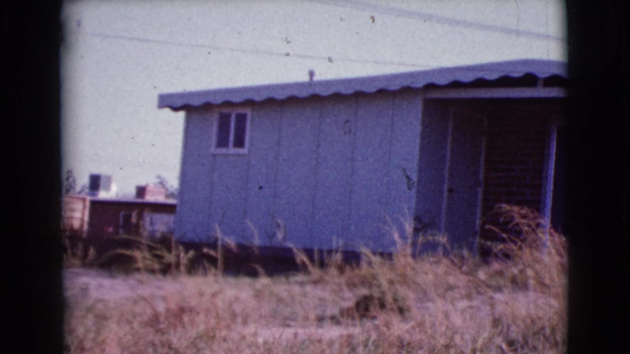 Light-colored mobile home behind dry grass with a pale sky and nearby structures.