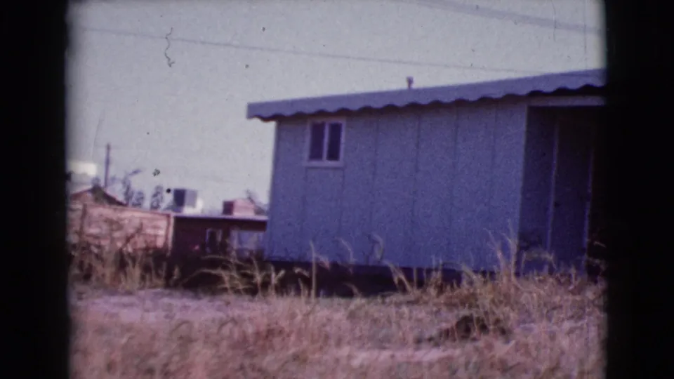 Cottonwood, Arizona home-movie frame showing a modest mobile home, dry overgrown grass, and simple neighboring structures.