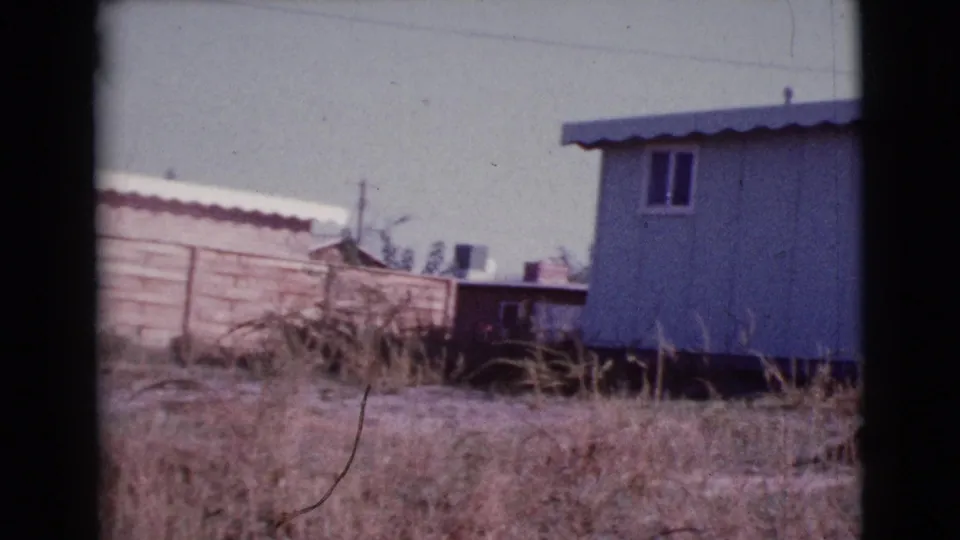 1964 8mm frame from Cottonwood showing a wooden fence, dry lot, background buildings, and the edge of a mobile home.