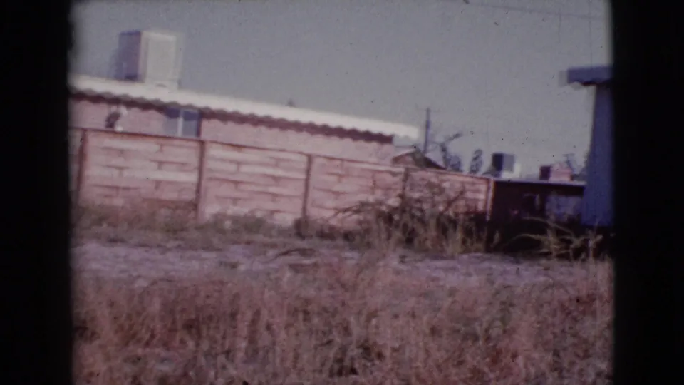 Cottonwood, Arizona frame showing a simple fence, dry summer grass, nearby rooflines, and the cropped edge of a mobile home.
