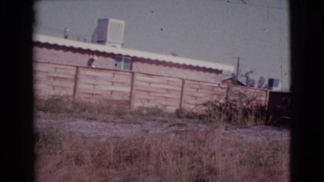 Low wall and white-roofed house behind dry grass, seen through dark 8mm frame borders.