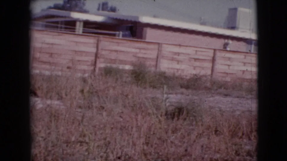 1964 8mm frame of a residential wall, white roof, shaded window area, and overgrown dry yard in Cottonwood, Arizona.