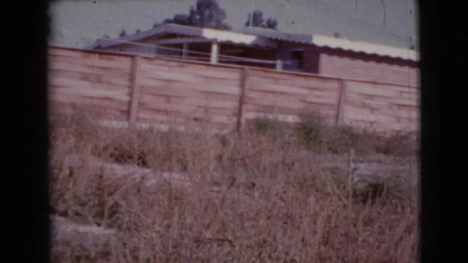 End frame from a 1964 Cottonwood, Arizona home movie showing a low wall, white roof, rooftop cooler, and dry yard.