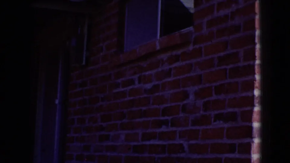 Dark, purple-tinged 8mm frame showing a brick wall and a narrow window or doorway edge at the end of the clip.