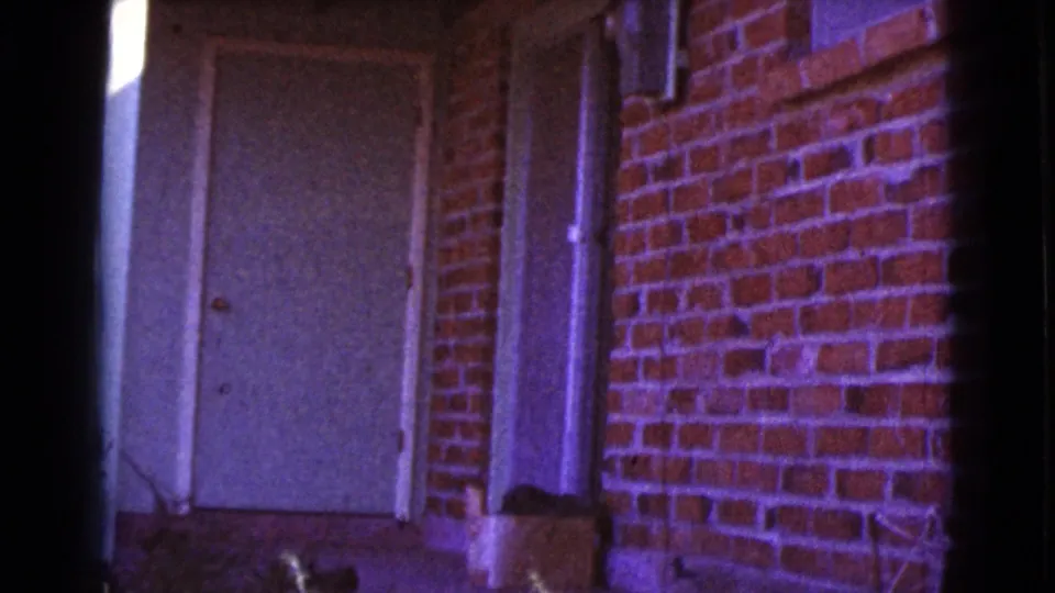 1964 Cottonwood, Arizona 8mm frame showing a white exterior door, red brick wall, pipe, and small box near the base of the building.
