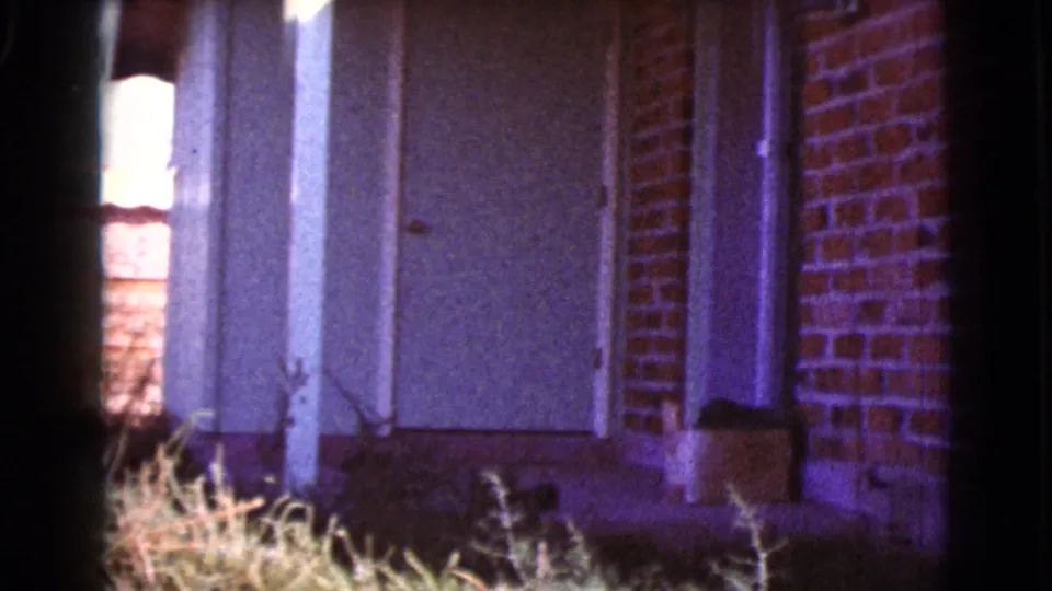 1964 Cottonwood, Arizona exterior frame showing a white door, red brick facade, utility pipe, and dry grass in the foreground.