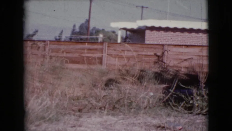 1964 Cottonwood, Arizona home-movie frame showing a wooden fence, dry grass, utility poles, wires, and a low building near distant terrain.