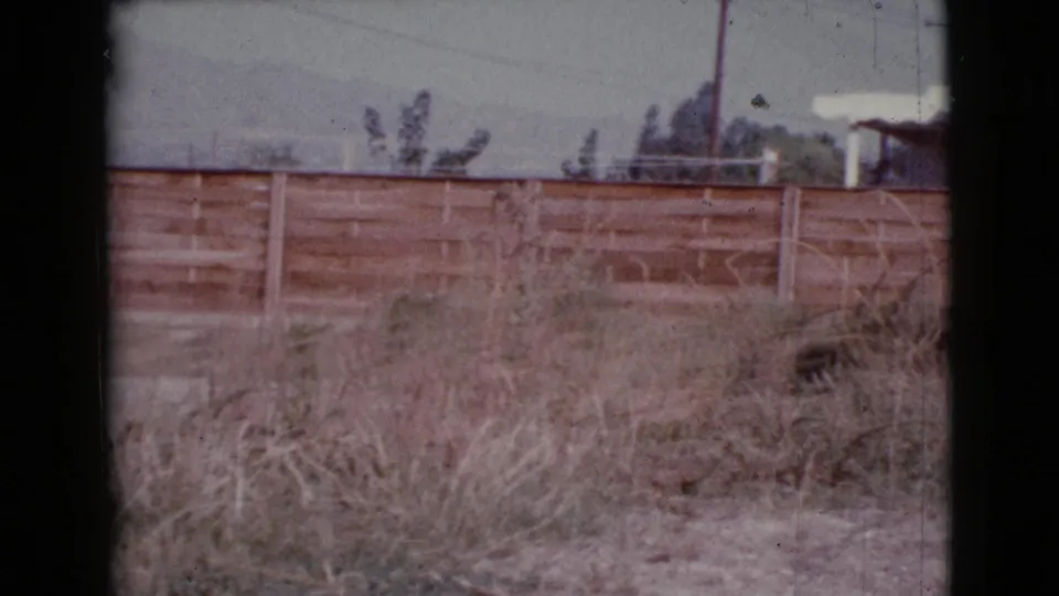 1964 Cottonwood, Arizona exterior frame with a long wooden fence, dry weeds, overhead wires, pale sky, and distant landscape forms.