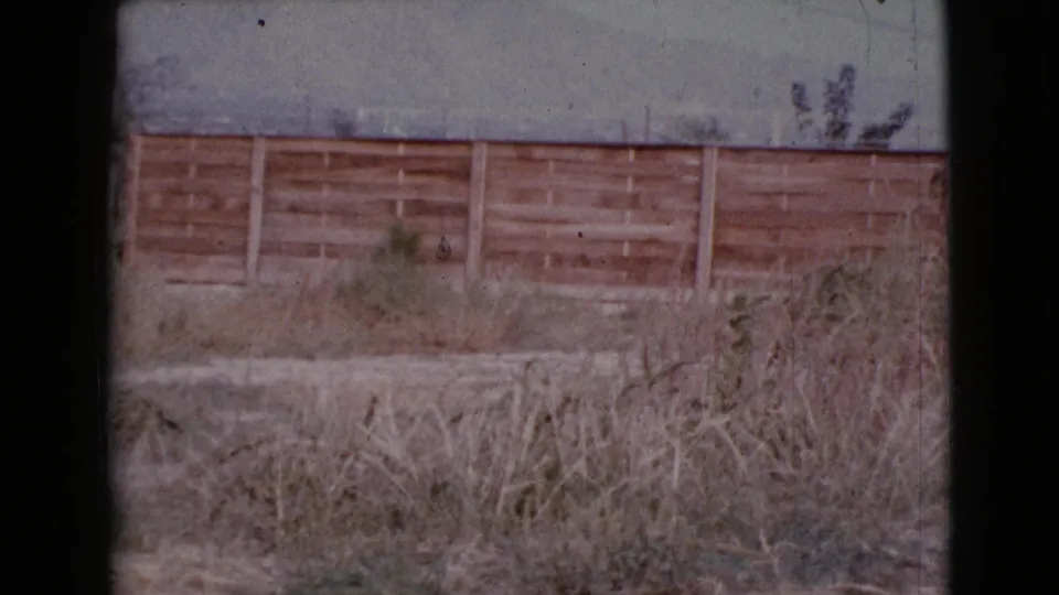1964 Cottonwood, Arizona home-movie frame with dry grass, a wooden fence, a low structure, utility lines, and a muted sky.