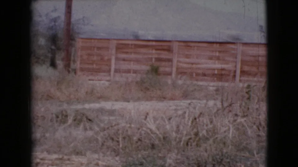 1964 Cottonwood, Arizona 8mm frame showing dry grass, bare ground, a wooden fence, and a utility pole under a hazy sky.