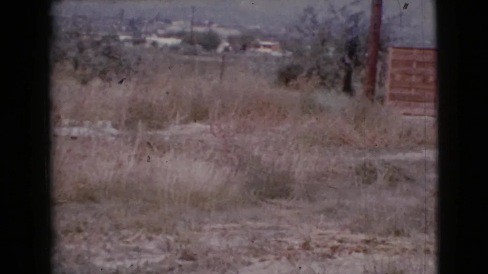 1964 Cottonwood, Arizona 8mm frame looking across dry grass and dirt toward distant buildings and hills, with part of a wooden fence at right.