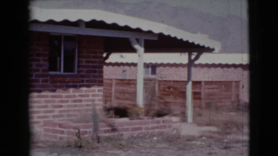 1964 Cottonwood, Arizona home-movie frame showing a red brick house with light corrugated roof, white porch posts, small window, and planter.
