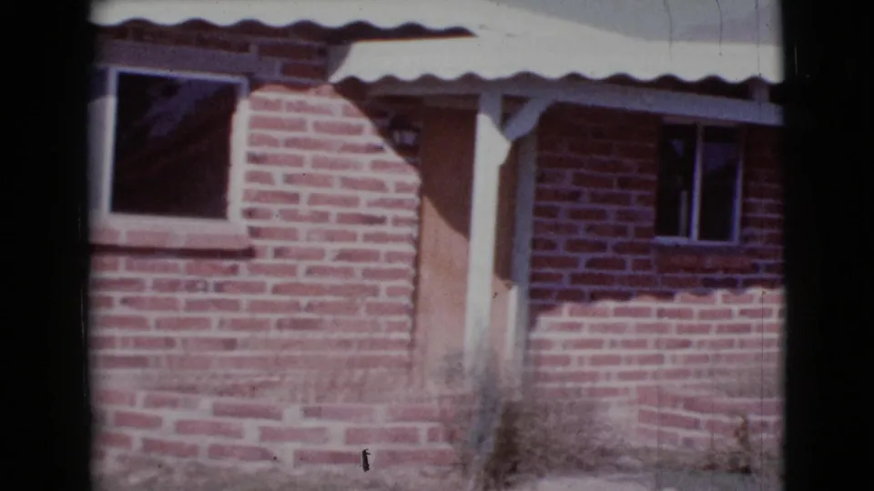 1964 Cottonwood, Arizona home-movie view closer to the red brick facade, showing a large window, doorway, roof edge, and porch posts.