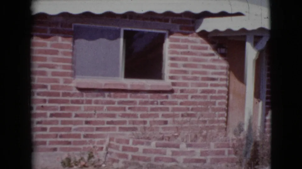 1964 Cottonwood, Arizona frame showing the brick house wall, large window, dark opening, porch edge, low planter, and sparse yard plants.