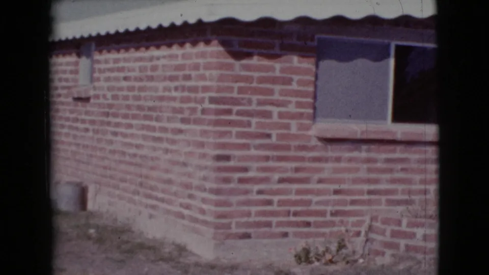 1964 Cottonwood, Arizona home-movie frame of a brick house corner with window, metal awning, concrete base, and dry yard.