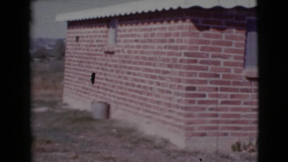 1964 Cottonwood, Arizona 8mm frame showing the side wall of a brick home, open yard, sparse plants, and distant terrain.