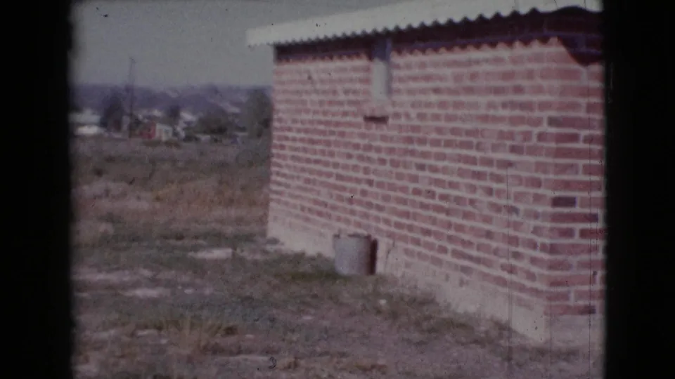 1964 Cottonwood, Arizona frame of a brick residential side wall, corrugated roof edge, dry yard, and low distant hills.