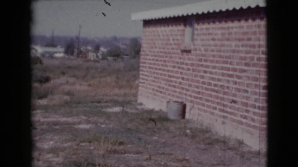 1964 Cottonwood, Arizona 8mm frame showing a brick exterior wall, dry field, and distant residential area under a pale sky.