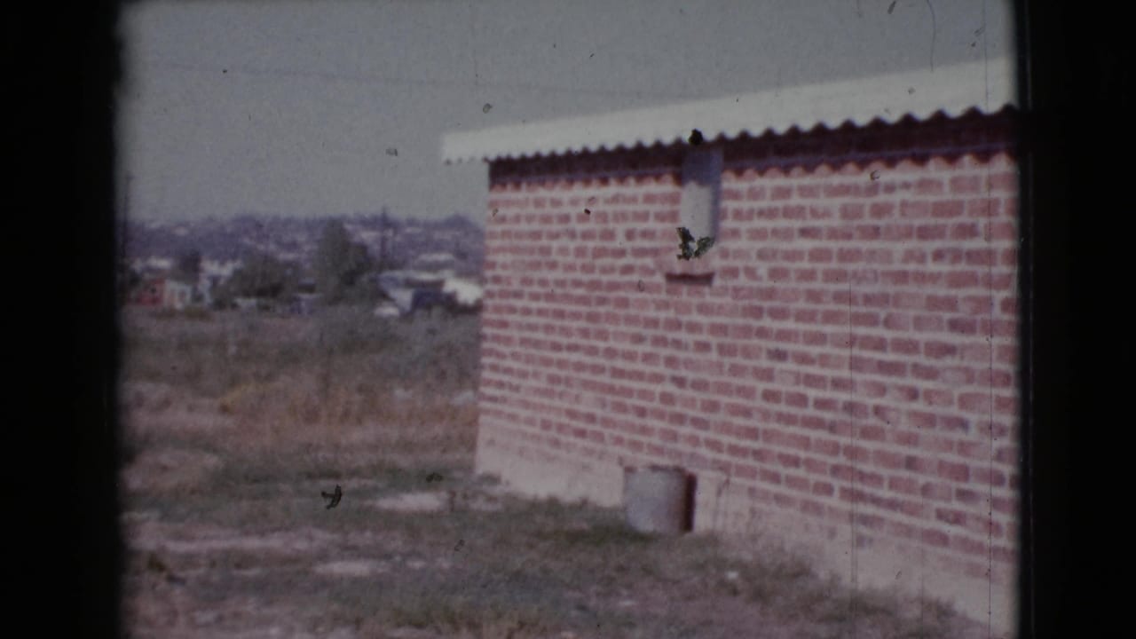 Red brick building wall with corrugated roof edge beside dry open ground.