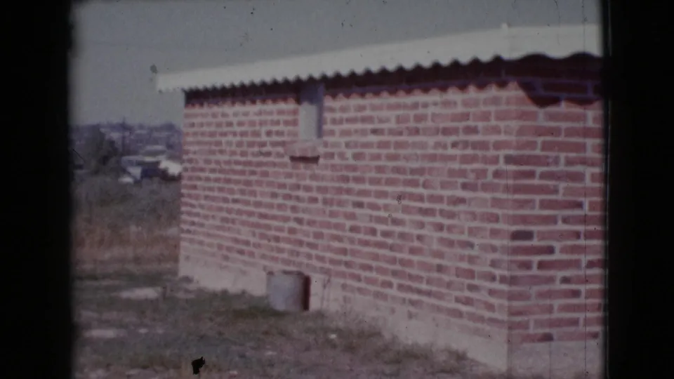 1964 Cottonwood, Arizona 8mm frame of a red brick house corner with a corrugated roof edge and sparse open ground nearby.