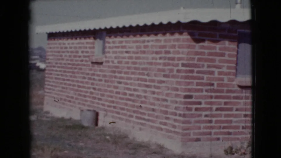1964 Cottonwood, Arizona 8mm frame looking along a red brick house wall with small openings, roof overhang, and dry grass at the base.