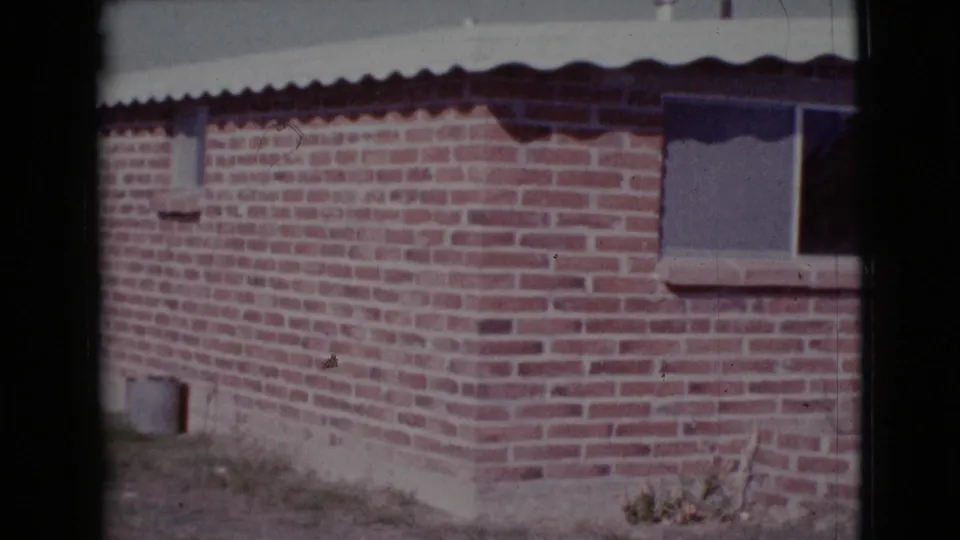 1964 Cottonwood, Arizona 8mm frame showing the side of a red brick house with a small window, roof overhang, and dry vegetation below.