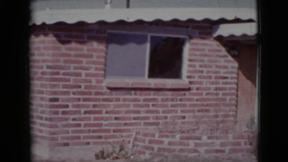 1964 Cottonwood, Arizona 8mm frame showing a brick house wall, window, and partial doorway area in bright sun.