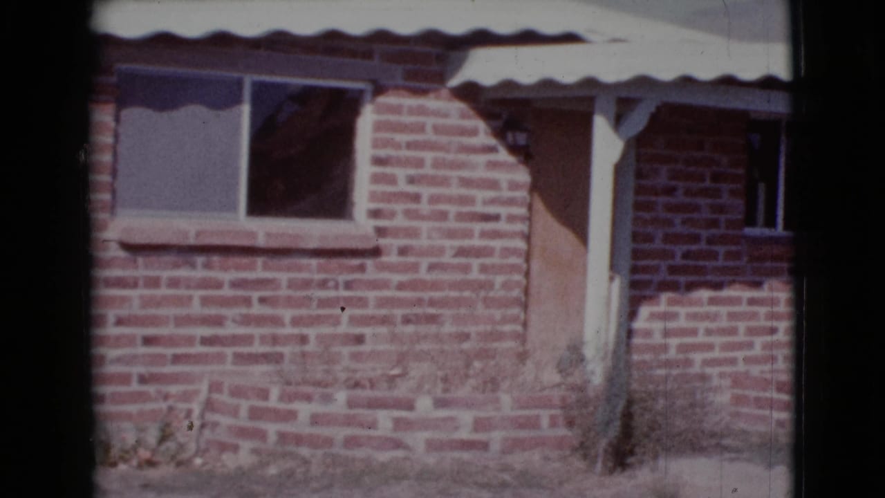 Brick house facade with a white corrugated awning, window, porch post, and boarded doorway.