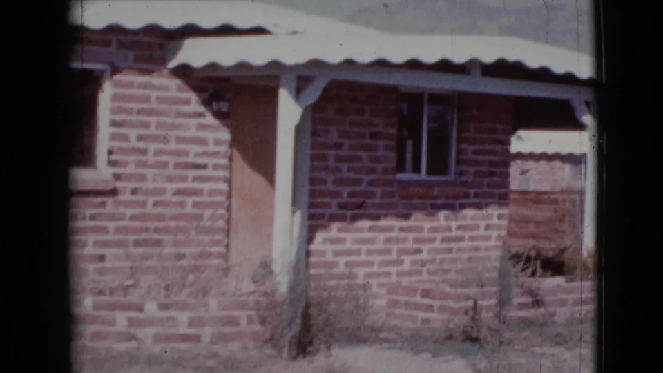 1964 Cottonwood, Arizona home-movie frame showing the brick exterior closer in, with porch post, awning edge, window, and low planting bed.