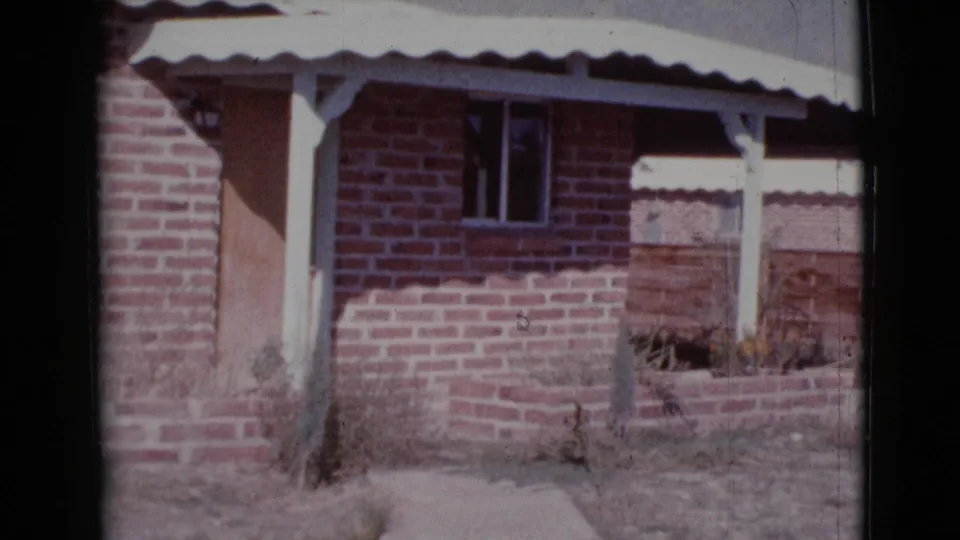 1964 Cottonwood, Arizona frame showing the brick house exterior from farther right, including porch posts, white awning, dark window, and bare ground.