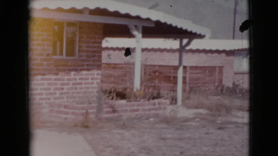 A faded 8mm frame shows the 1964 Cottonwood brick home exterior with white awning, shaded porch, low planter, and sparse vegetation.