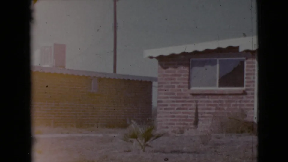 The final frame pans to another section of the Cottonwood residence, showing a brick wall, window, low adjacent structure, dry yard, and small plant.