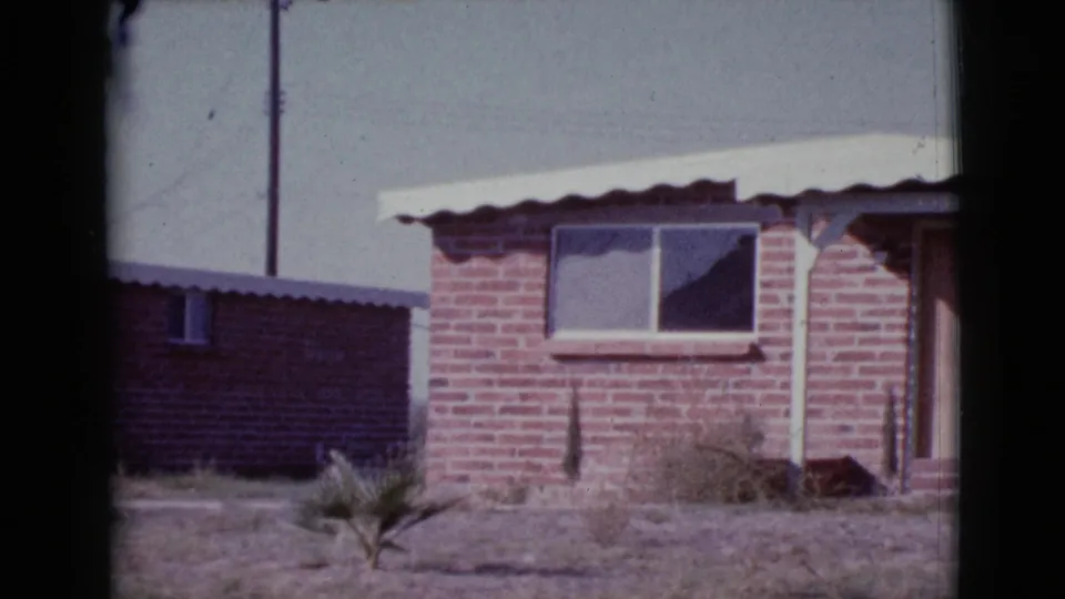 1964 Cottonwood, Arizona 8mm frame showing the brick house corner, shaded window, dry yard plant, and partial entry area.