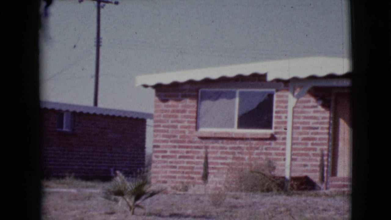 Brick house exterior with corrugated roof, small window, dry yard, and dark film border.