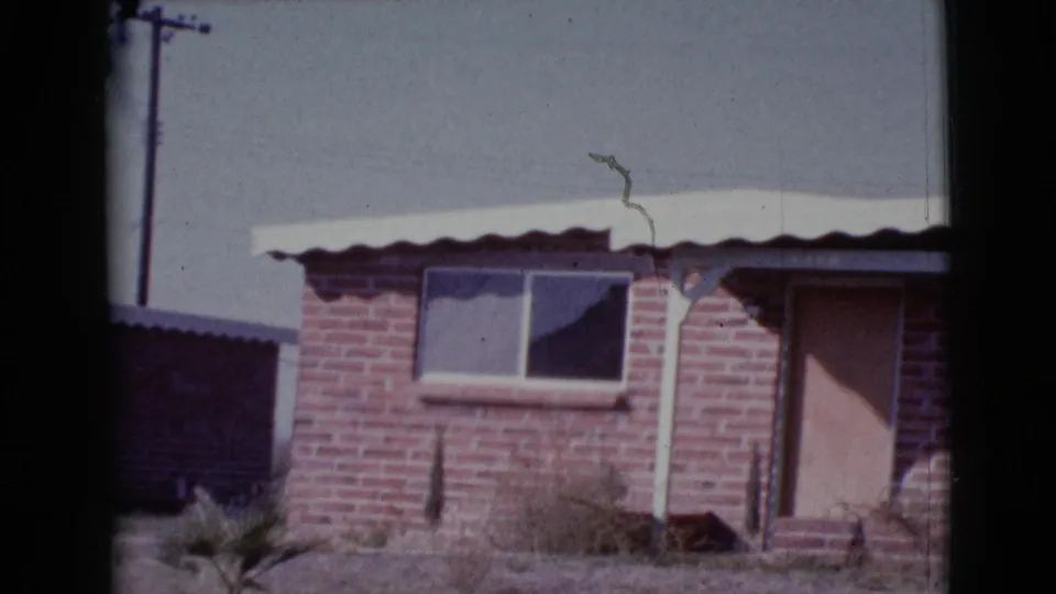 1964 Cottonwood, Arizona 8mm frame of the brick house facade, showing a small window, pale metal roof, boarded door, and desert yard.