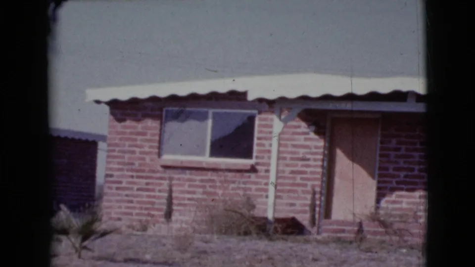 1964 Cottonwood, Arizona 8mm frame showing the house front with brickwork, small window, boarded entry, roof post, and sparse desert plants.