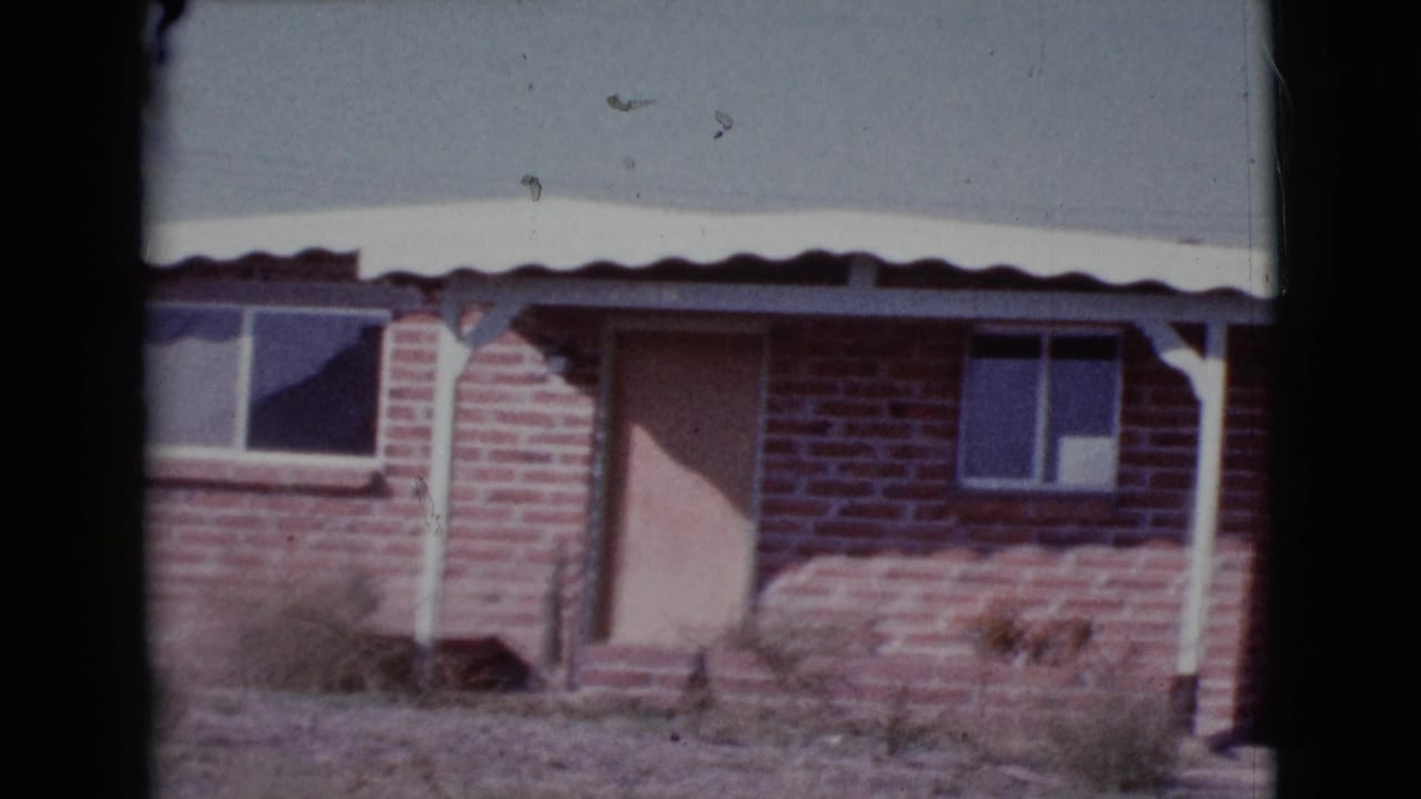 Brick house exterior with pale door, small windows, tin roof edge, white posts, and dry weeds.