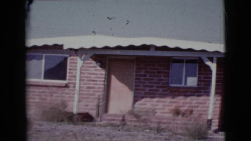 1964 Cottonwood, Arizona 8mm frame showing a red brick house facade with a pale door, small windows, tin roof edge, white posts, and weeds.