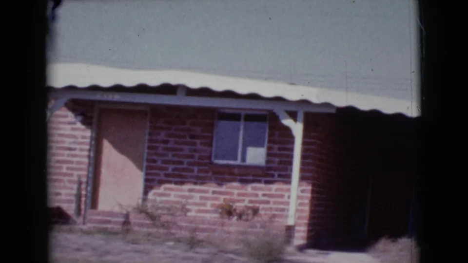 1964 Cottonwood, Arizona frame of a weathered brick house exterior with a door, window, white post, scalloped roof edge, weeds, and shadow.
