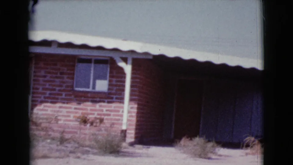 1964 Cottonwood, Arizona 8mm frame showing the pan farther along the brick house, with a window, white post, dark recess, roof edge, and weeds.