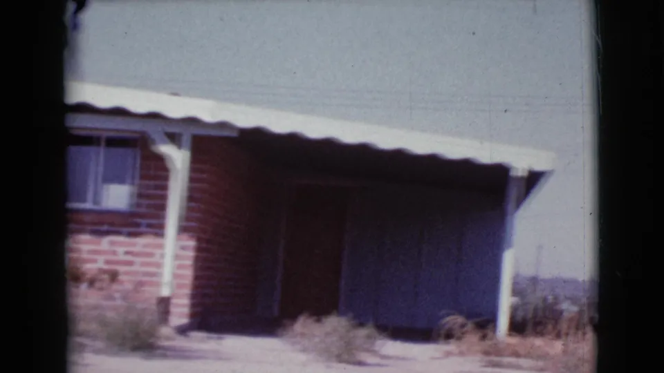 1964 Cottonwood, Arizona frame at the end of the pan, showing the brick house corner, white posts, dark recessed openings, roof edge, weeds, and bright sky.