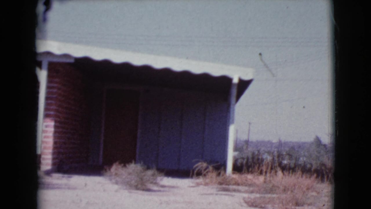 Brick house exterior with a dark garage or carport opening, white awning, dry grass, and concrete drive.