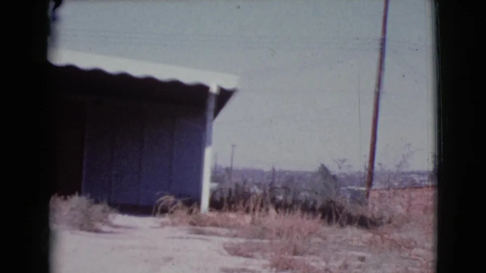 1964 Cottonwood, Arizona frame with the house edge at left, a dark garage or carport opening, dry yard, utility pole, and distant rooftops.
