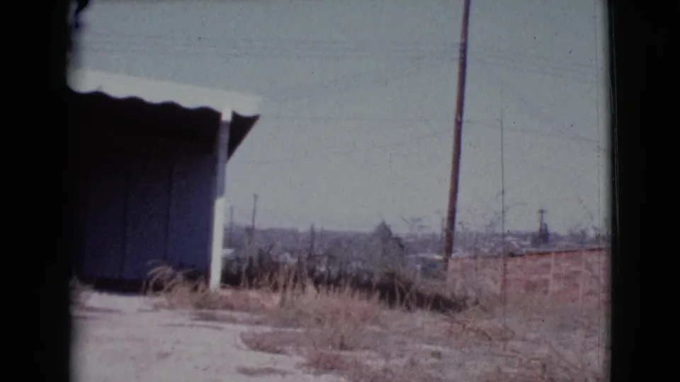 1964 Cottonwood, Arizona 8mm frame showing a partial house edge, dry residential yard, utility pole, low wall, and hazy distant neighborhood.