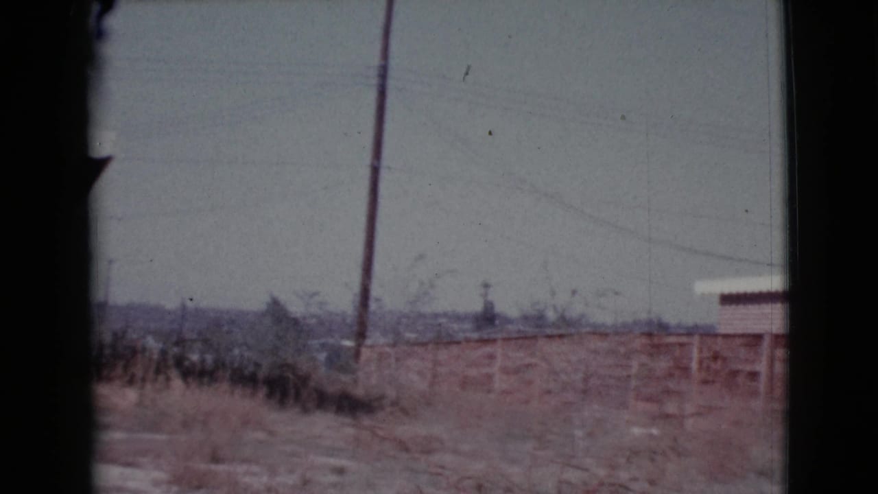 Dry field with utility pole, low fence, distant town structures, and hazy sky.