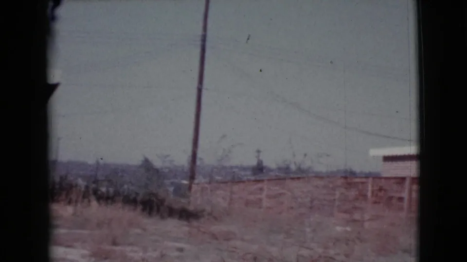 1964 Cottonwood, Arizona home-movie frame looking across a dry field toward low distant buildings and a utility pole.